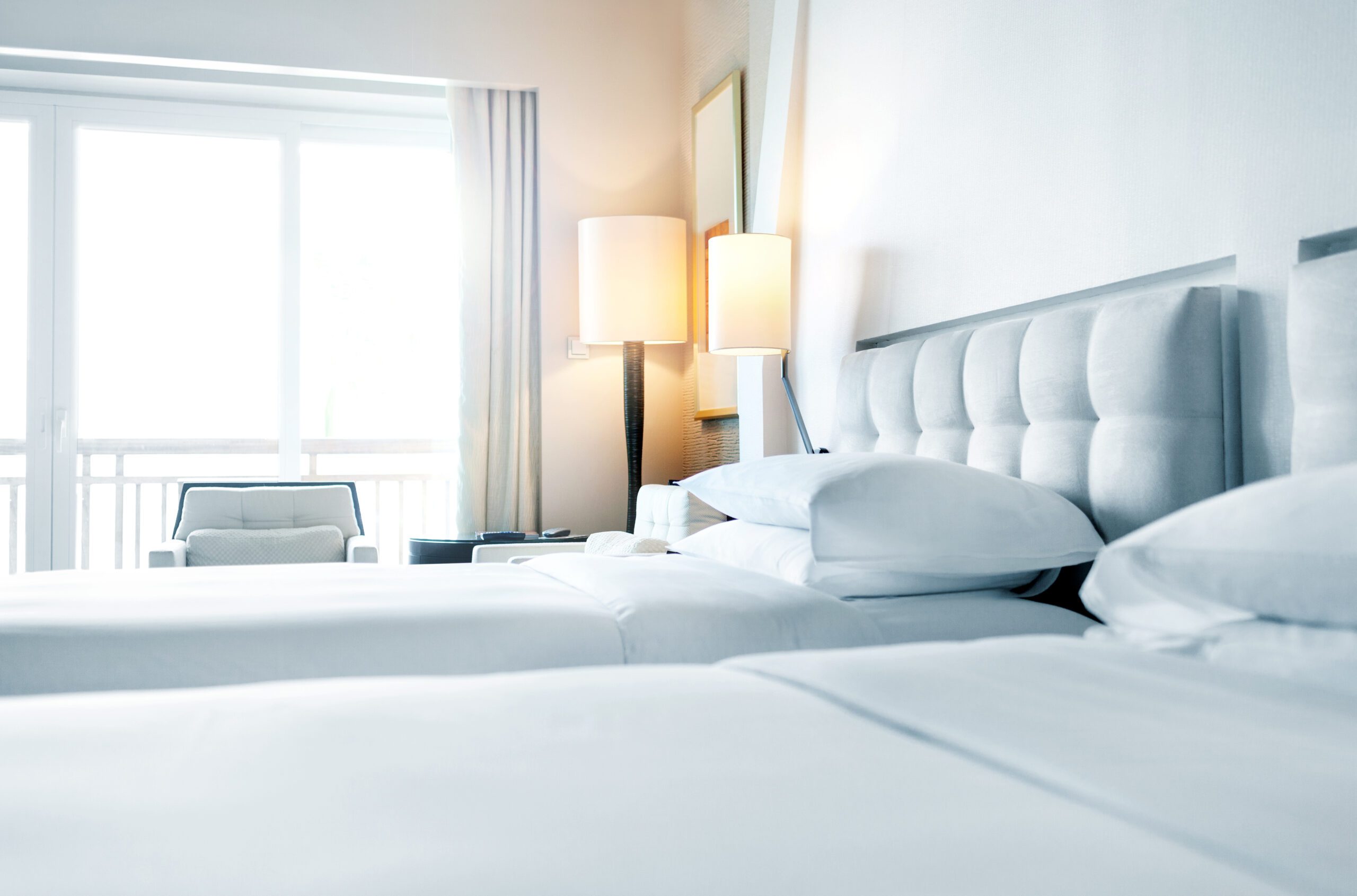 Two twin-sized white linen beds with white headboards and a lit up lamp.