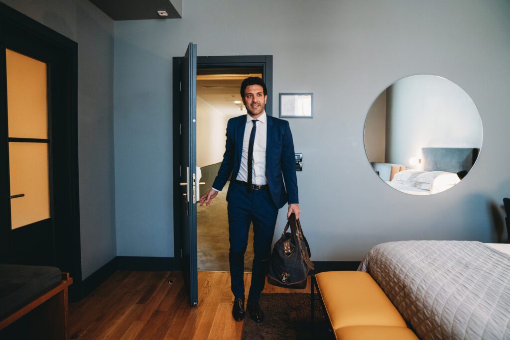 Businessman in blue suit with a leather bag entering a hotel room. Wood flooring, With a white and grey bed and orange cushions.